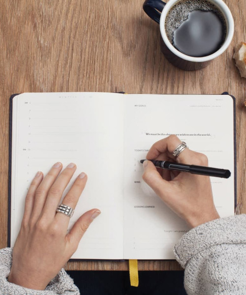A table with a notebook, a coffee mug and a croissant from above. A women is taking notes.