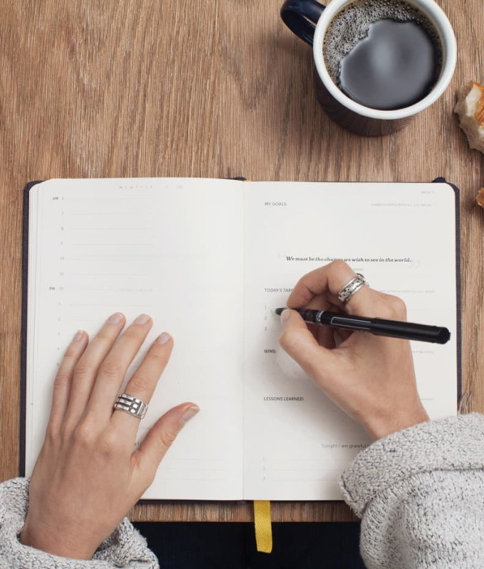 A table with a notebook, a coffee mug and a croissant from above. A women is taking notes.